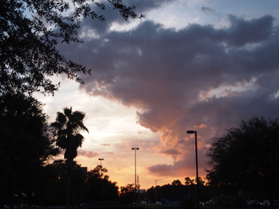 [A large thick bank of clouds covers the upper right half of the sky. Behind this cloud to the left and top are light blue skies. The lower levels of this cloud have a light-orange tinge. The light posts and trees in the image are dark because the sky behind them is so bright.]